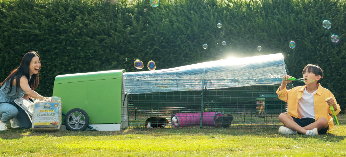 Un enfant fait des bulles dans le jardin, tandis que sa mère change le foin dans le clapier de Eglu Go .