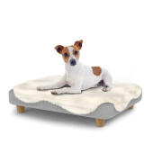 Chien assis sur un petit lit pour chien Topology avec une couverture en peau de mouton et des pieds ronds en bois.