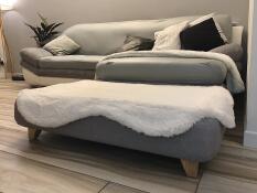 Un lit gris avec un surmatelas blanc en peau de mouton et des pieds carrés en bois